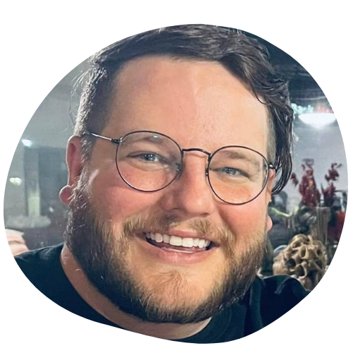 A smiling man with short brown hair, a beard, and round glasses is shown in close-up. He is wearing a black shirt, and there are blurred indoor decorations in the background.